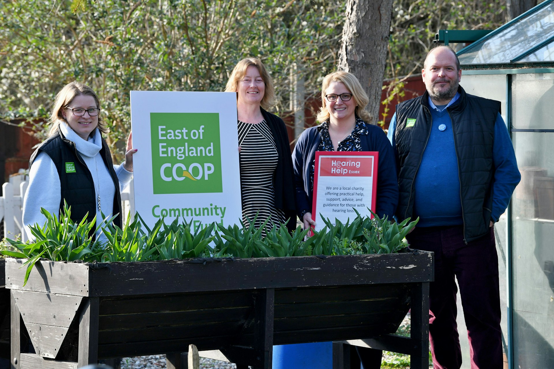 Four people standing beside a planter holding East of England Co-op and Hearing Help Essex signs